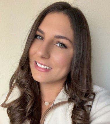 Portrait of a smiling woman with long brown hair and blue eyes, wearing a light beige top against a pale wall.